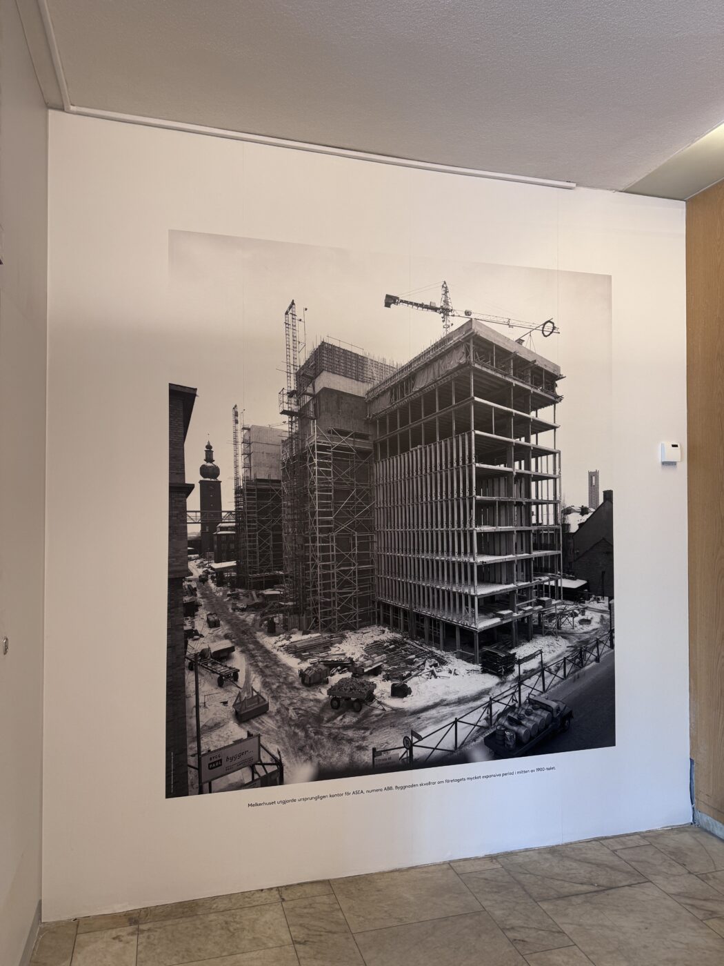 Large black-and-white photo of a building under construction with cranes, mounted on a white wall in a gallery setting (indoor display).