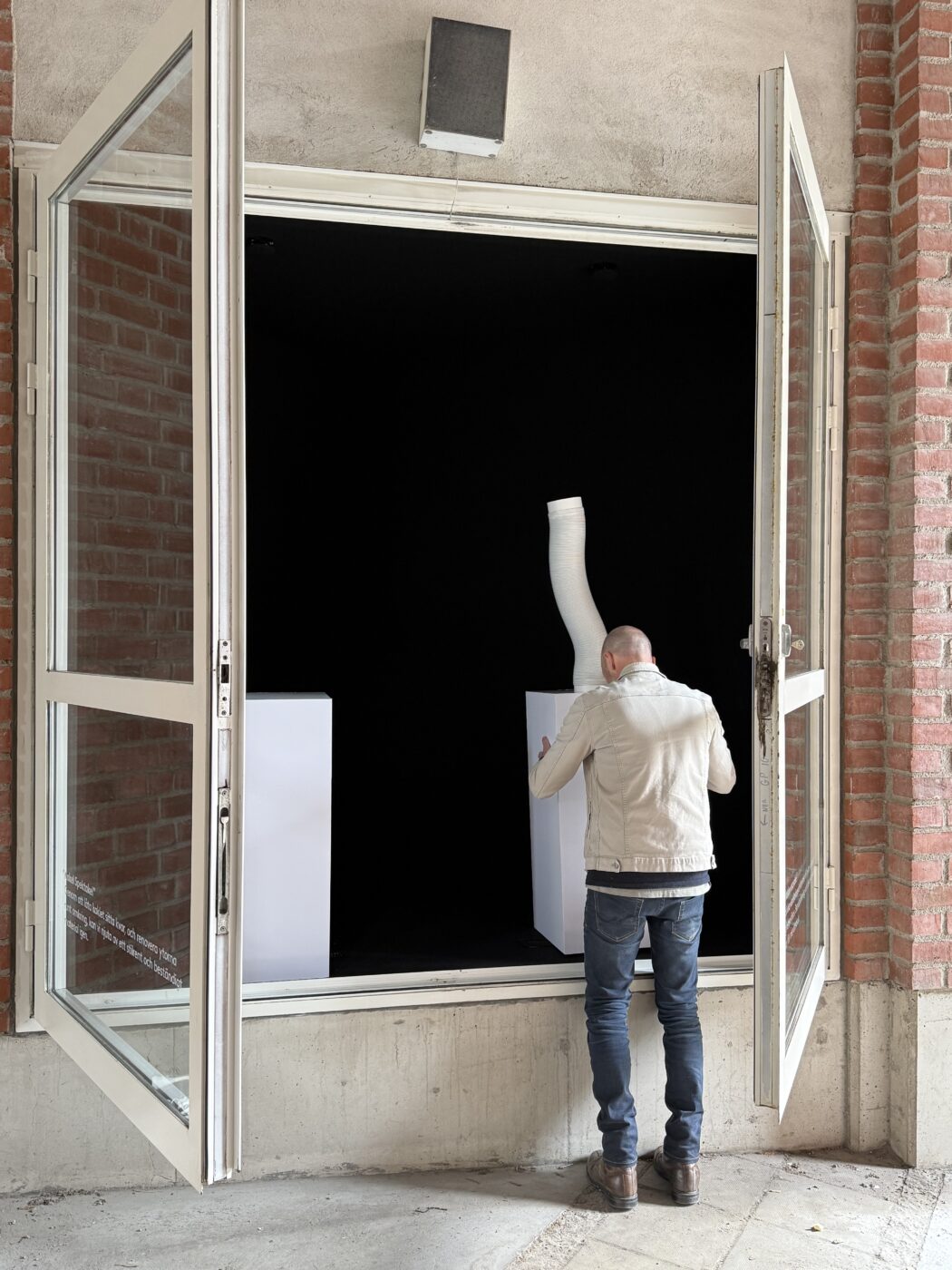 Man in a beige jacket carries a white panel through an open glass storefront door into a dark interior.