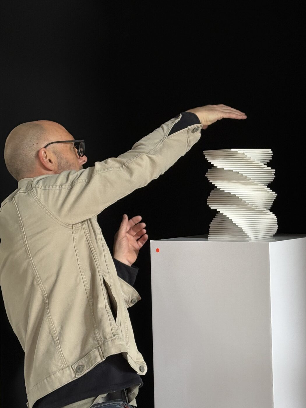 Man in a beige jacket balances a tall spiral stack of flat white tiles on a pedestal against a dark background.
