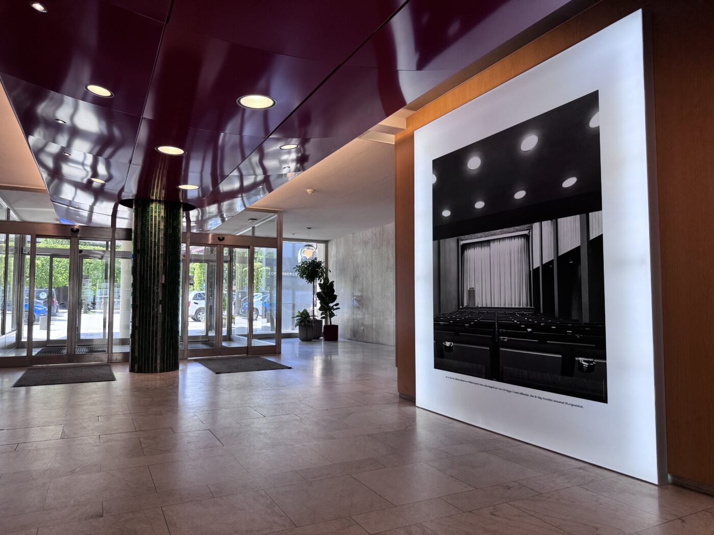 Lobby interior with a glossy purple ceiling, circular recessed lights, glass entrance doors, and a large framed black-and-white photo on the right wall.