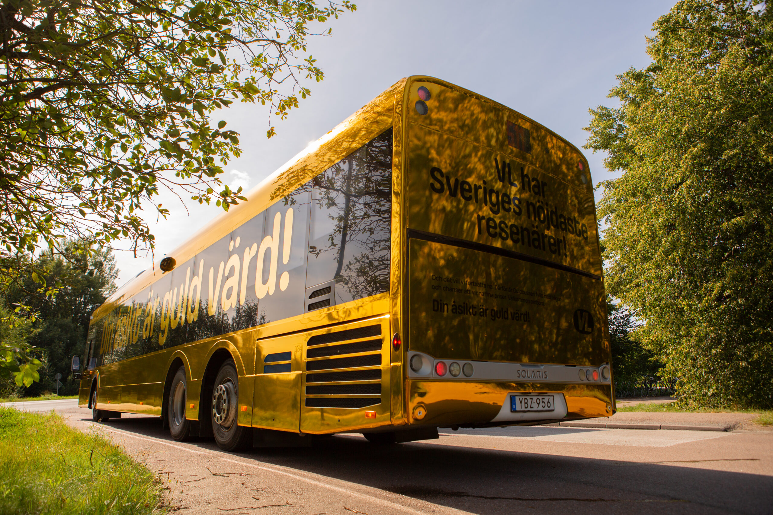 Golden bus parked on a road, its shiny yellow surface reflecting sunlight with trees around it.