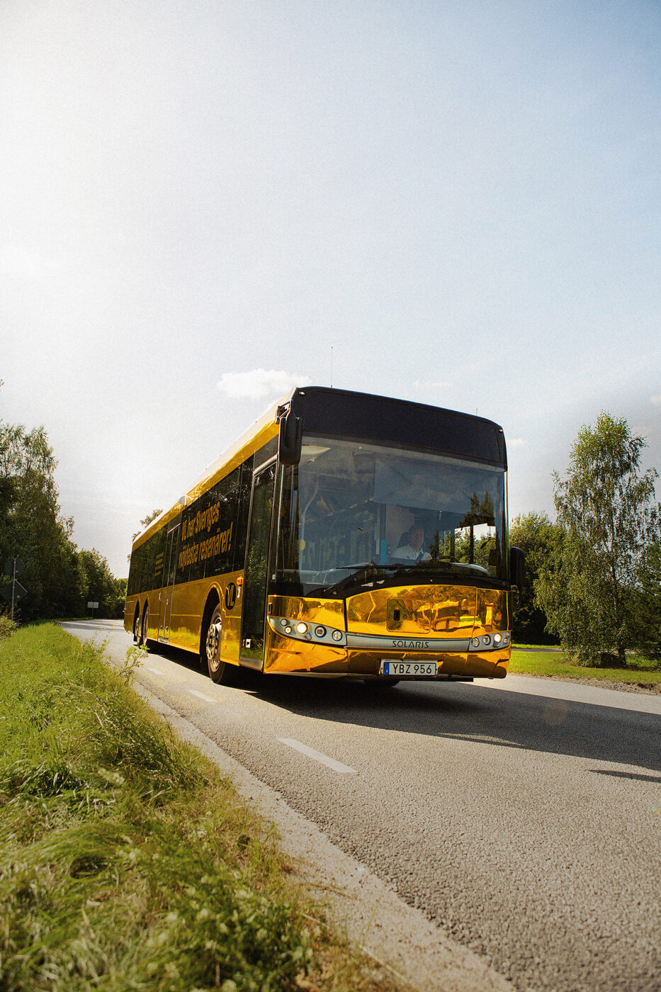 Yellow and black bus driving along a grassy, tree-lined rural road on a sunny day (front three-quarter view).
