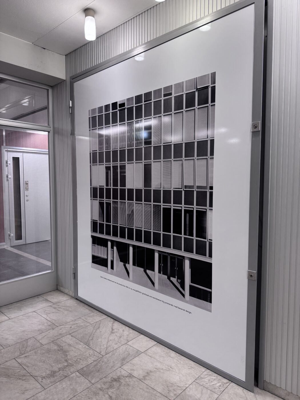 Large framed poster showing a grid of square tiles, mounted on a metal-framed wall in a corridor with tiled floor