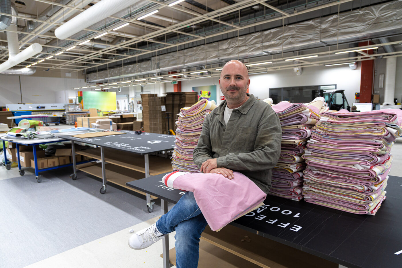 Man sits at a table with a tall stack of folded pink textiles in a workshop or factory setting behind him.