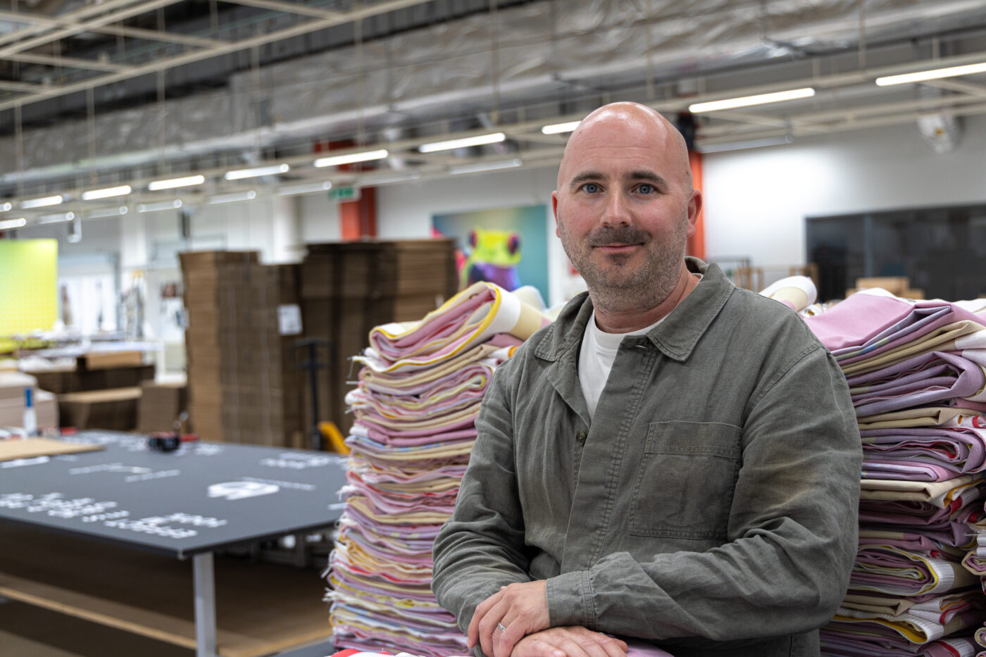 Smiling man standing beside stacked folded fabrics in a textile warehouse theater-like setting.