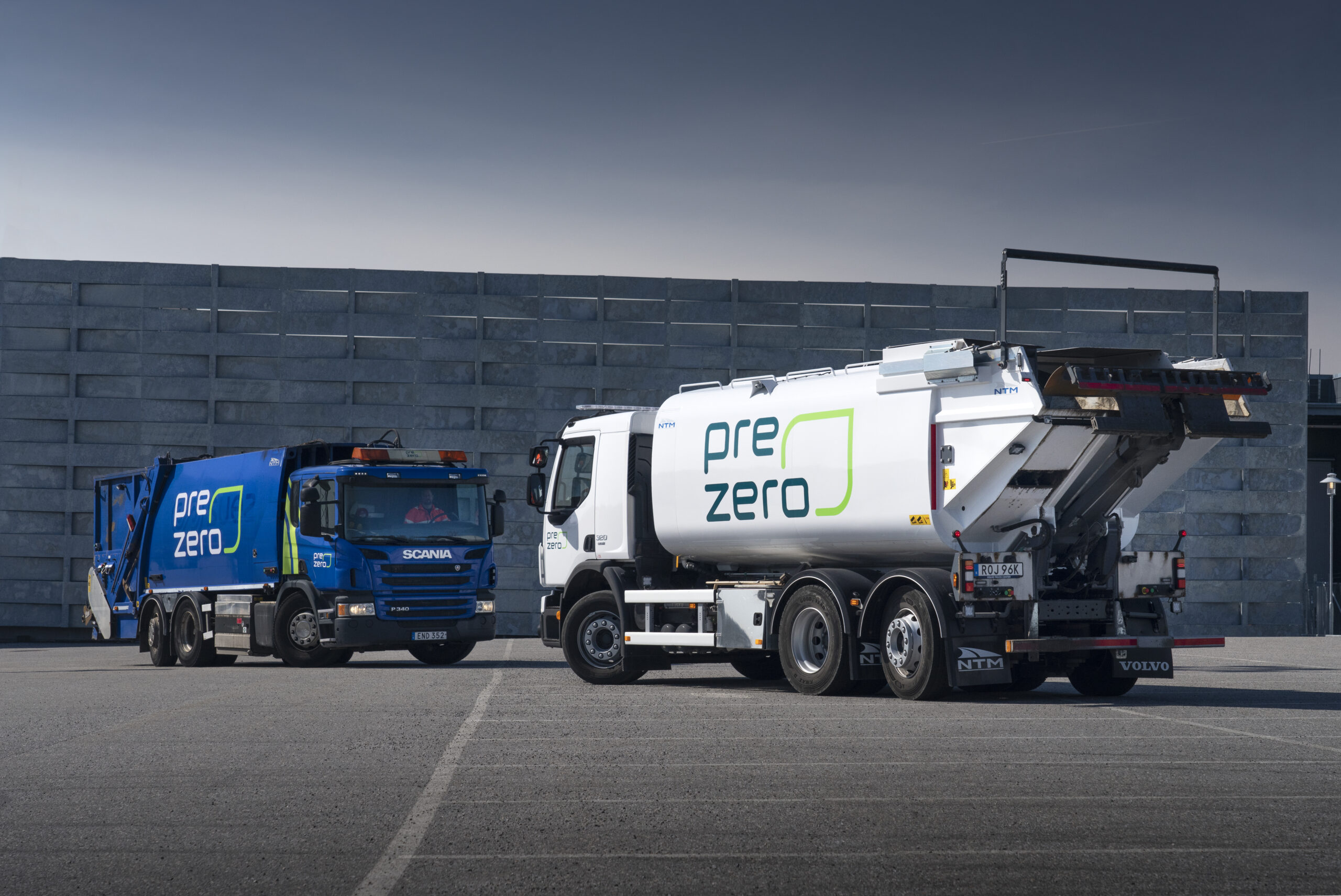 White tanker truck with green PreZero logo beside a blue truck in a paved lot with a gray concrete wall behind.
