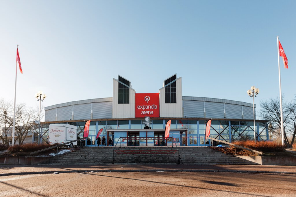 Expanda Arena building with a red central sign, glass entrance, and flags on both sides, set against a clear blue sky.