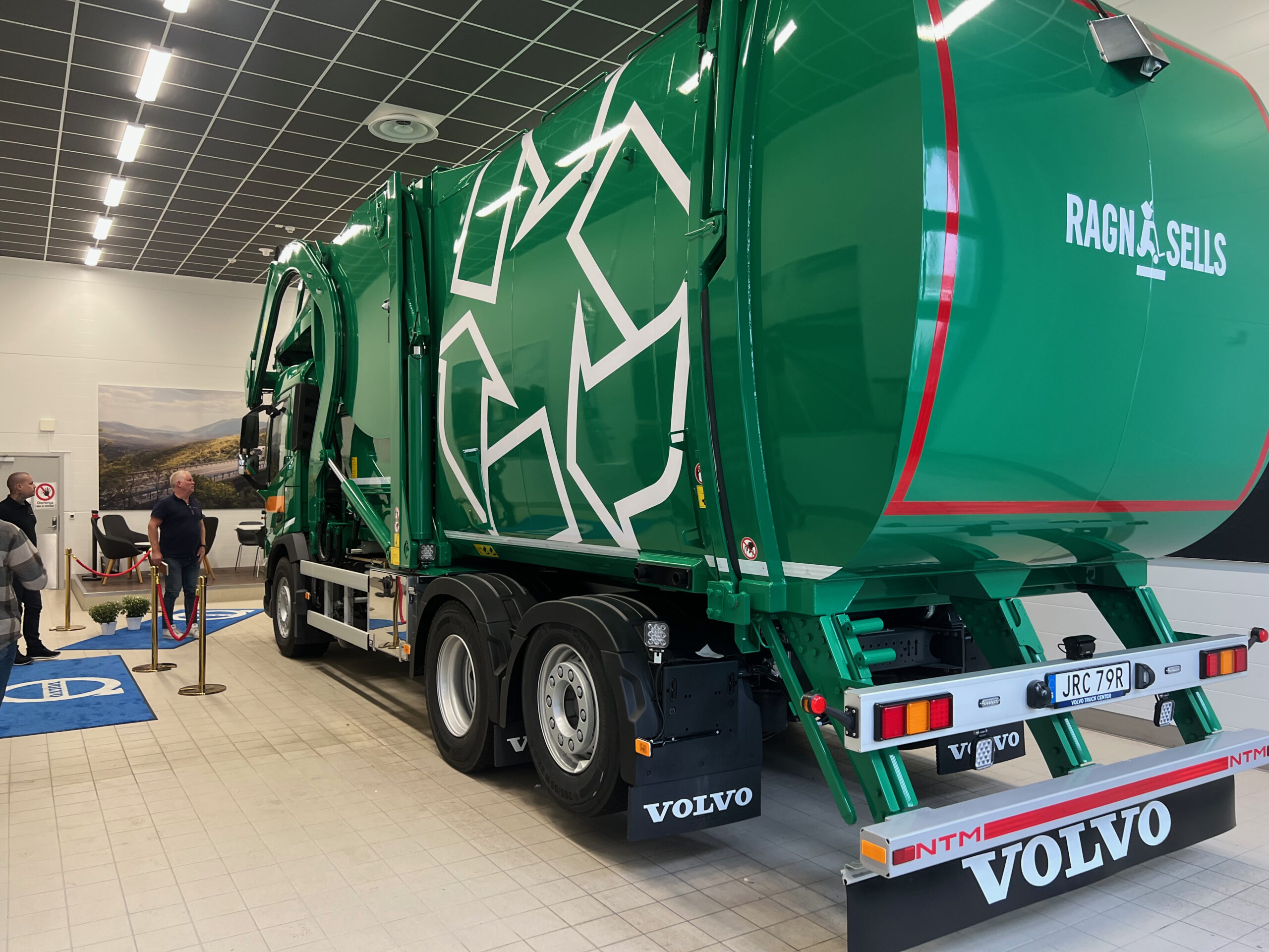 Green Volvo truck on display in showroom with attendants nearby and blue floor markings visible in foreground.