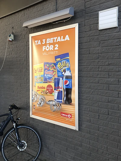 Poster on a dark brick wall advertising snacks and drinks (cola, chips) with a bicycle parked nearby; text in Swedish reads 'TA 3 BETALA FÖR? VÄLJ FRITTI!'